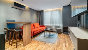 A compact living space with a sofa, a small dining table, a bar stool, a TV mounted on the wall, and a large window with curtains. - Urban Quarters - Gülhane (Istanbul)