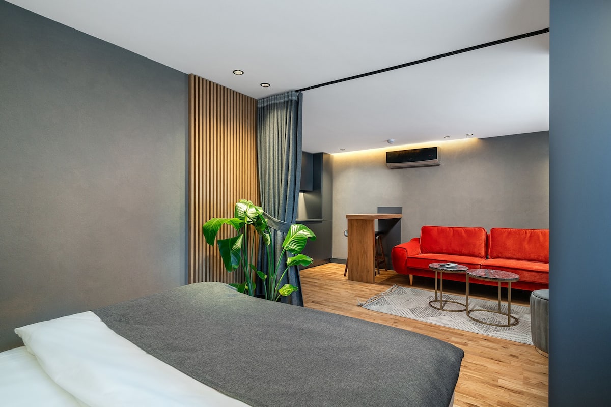 A modern hotel room with a bed, a red sofa, two small tables, a large plant, and a wooden floor.