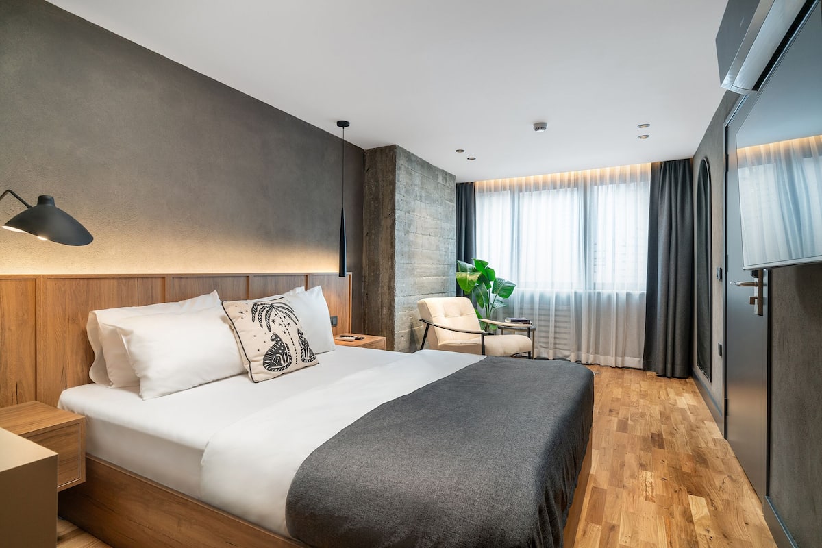 A modern hotel room with a large bed, a desk, a chair, and a plant.