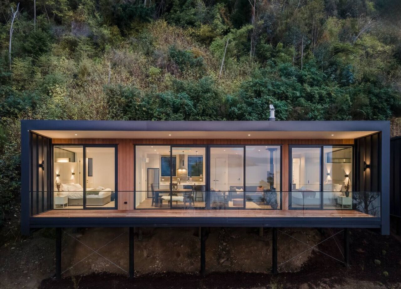 A modern, glass-enclosed cabin with a wooden deck, surrounded by forest.