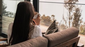 A person sitting on a wooden bench with a view of a lake and mountains through large windows.
