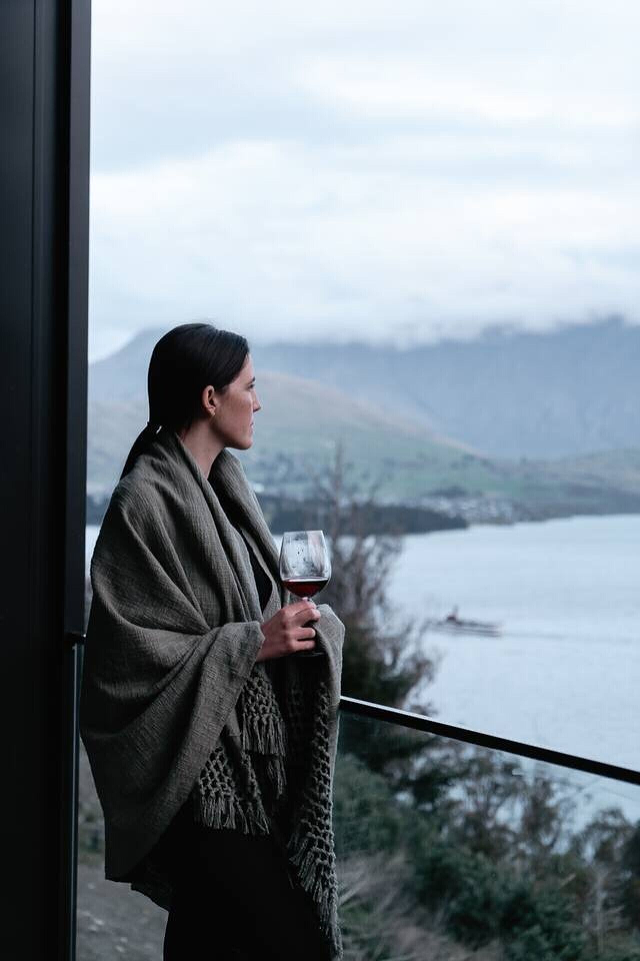 Une personne se tient près d’une fenêtre, un verre de vin rouge à la main, et on aperçoit un lac et des montagnes au loin.