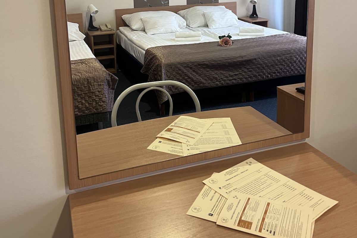 A hotel room with a bed, bedside tables, a desk with papers, and a mirror reflecting the room.
