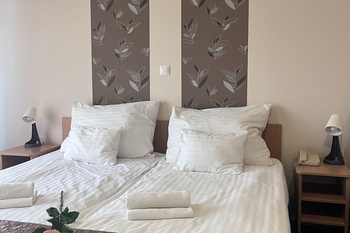 A neatly made bed with white linens, a brown quilt, and a single rose on the bed. There are two bedside tables with lamps and a telephone on one of them. The walls have a leaf pattern wallpaper.