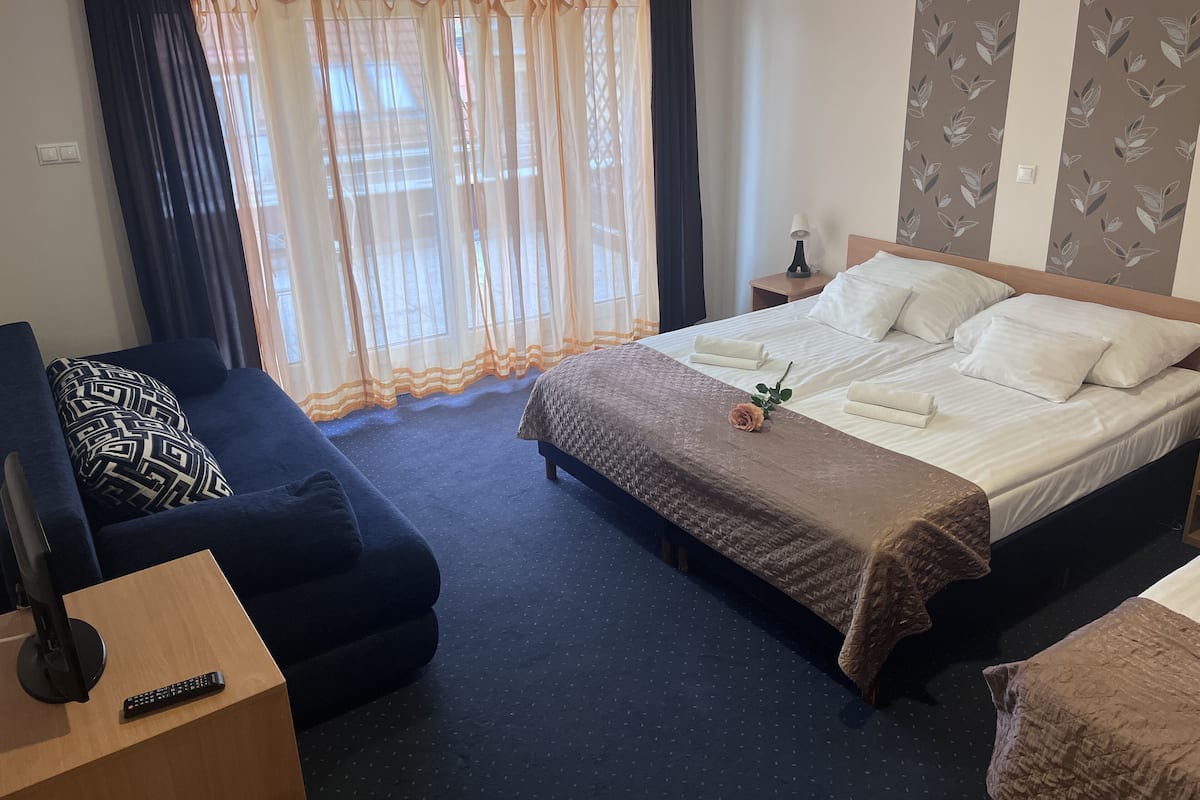 A hotel room with a bed, a sofa, a TV, and a remote control on a side table.