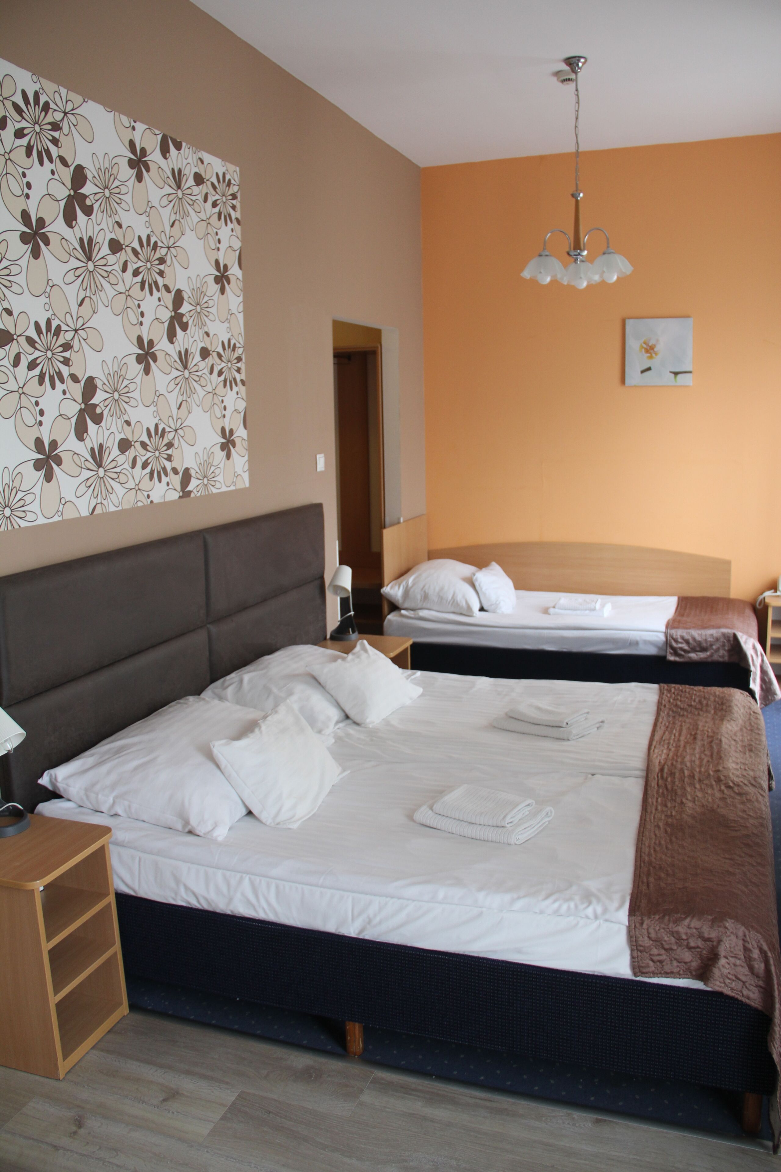 A hotel room with two beds, a wooden headboard, a nightstand, a lamp, and a floral wall decoration.