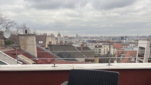 A balcony with a wicker chair, a table with a rose, and a view of a cityscape. - Hotel Rose City  (Budapest)