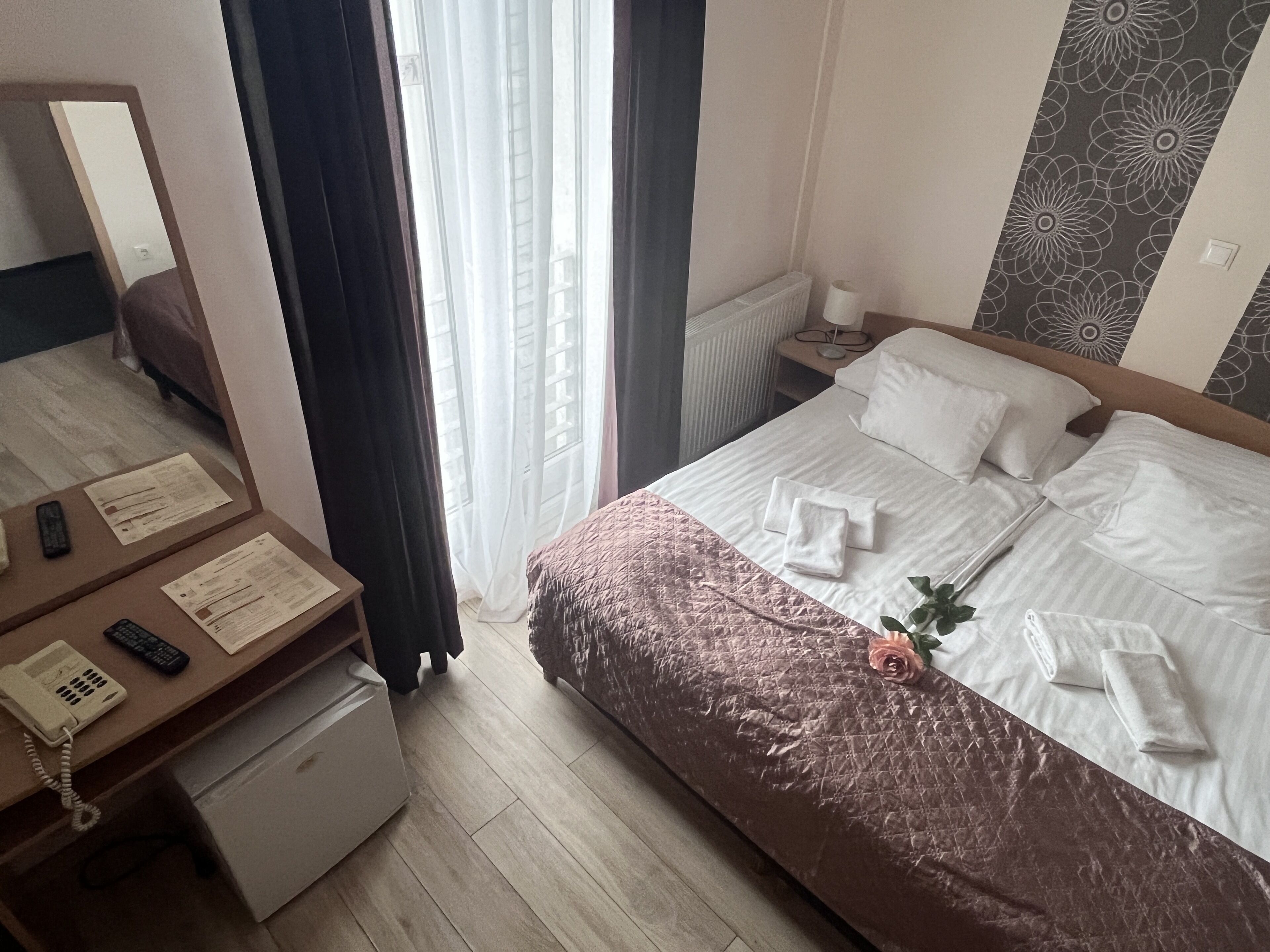 Hotellihuone, jossa on sänky, työpöytä, jossa on puhelin ja papereita, peili ja ikkuna, jossa on verhot.