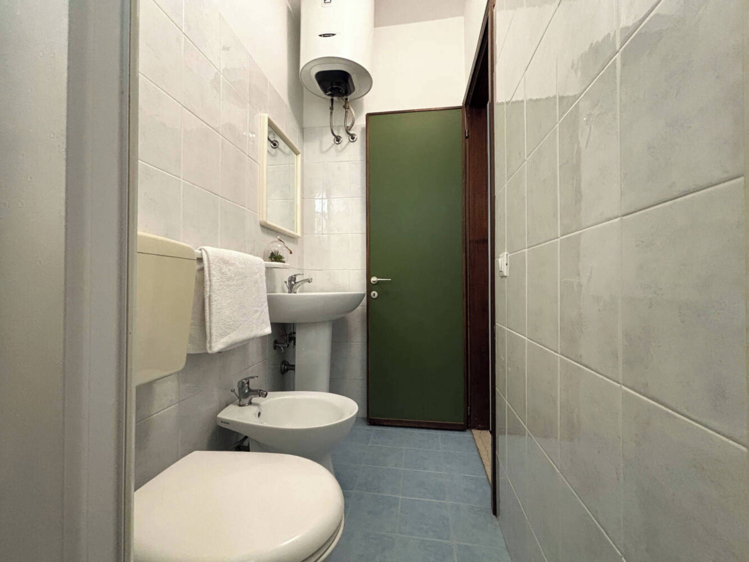 A small bathroom with a toilet, bidet, and sink. There is a green door and a white towel hanging on the wall.