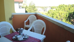 Ein Balkon mit Tisch für zwei Personen, weißen Stühlen und Blick auf Bäume und Gebäude im Hintergrund.