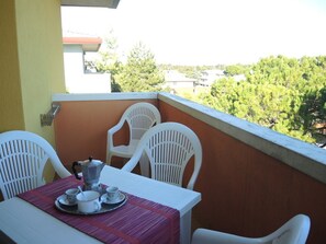 A balcony with a table set for two, white chairs, and a view of trees and buildings in the background.