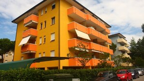 Ein mehrstöckiges, gelbes Gebäude mit orangefarbenen Balkonen, umgeben von parkenden Autos und Grünflächen.