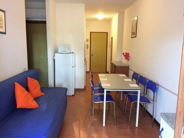 A compact living space with a blue sofa, a dining table with chairs, a refrigerator, and a small kitchenette. - Nice three-bedroom flat with balcony in Bibione (Bibione)