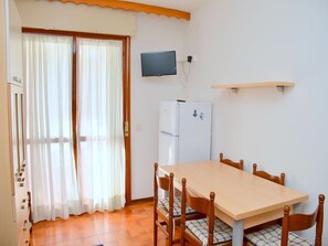 A dining area with a table and chairs, a refrigerator, a television mounted on the wall, and a sliding glass door with curtains. - Cozy two-room flat 100mt from Bibione beach (Bibione)