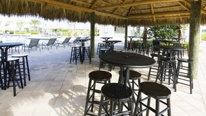Un'area patio esterna con tetto di paglia, diversi tavoli e sedie, e una piscina sullo sfondo.
