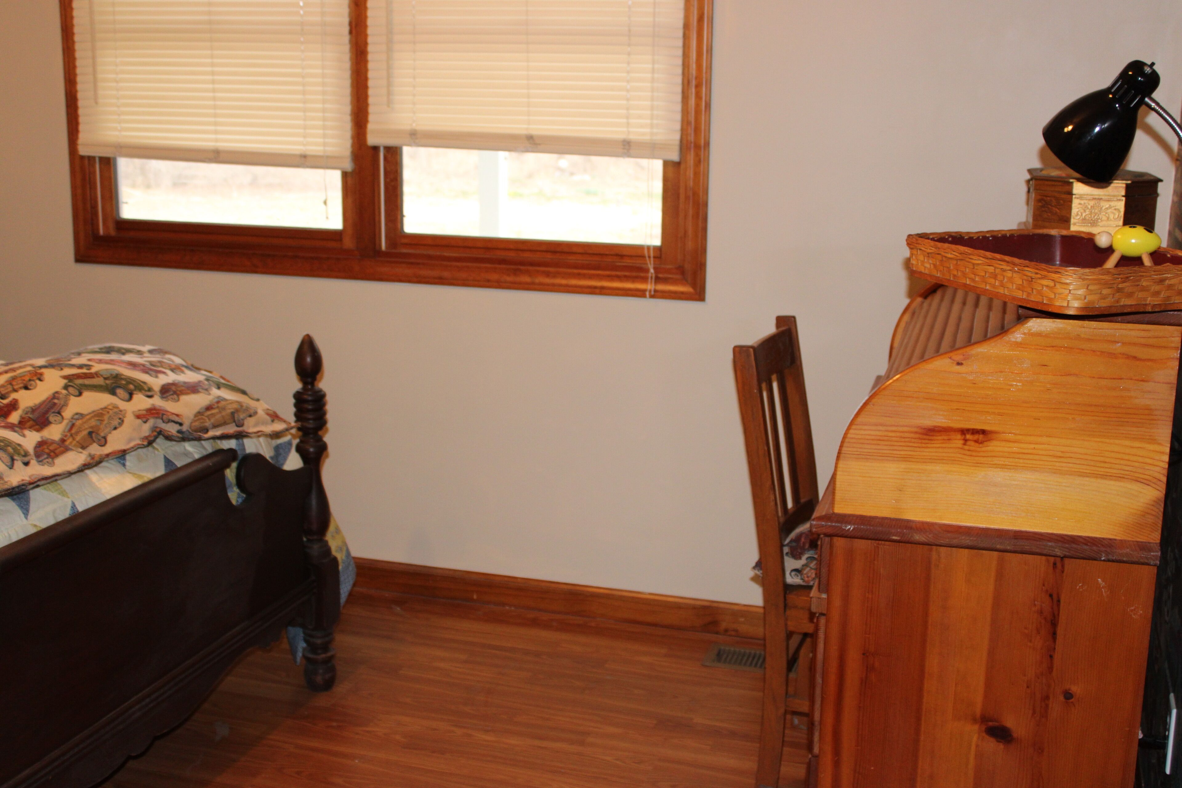 A bedroom with a wooden bed, a desk with a lamp, and a window with blinds.