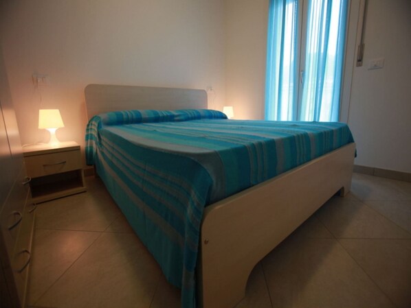 A bedroom with a bed, a nightstand, a lamp, and a window with curtains. - Elegant apartment near the beach (Rosolina Mare)