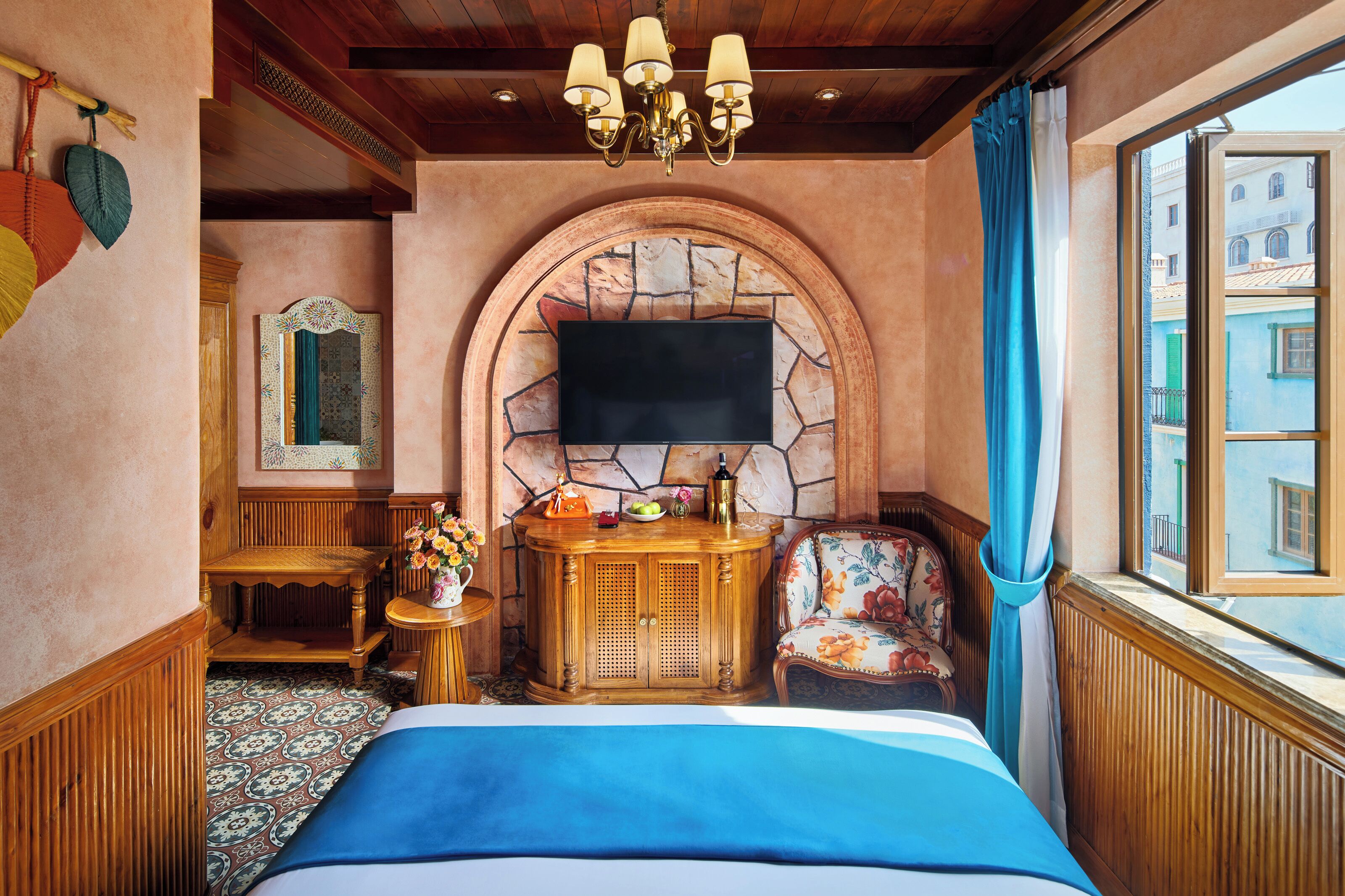 Una habitación de hotel con chimenea de piedra, un televisor de pantalla plana, una cama con cubrecama azul, una mesita de madera con un jarrón de flores y vista a edificios a través de una ventana.