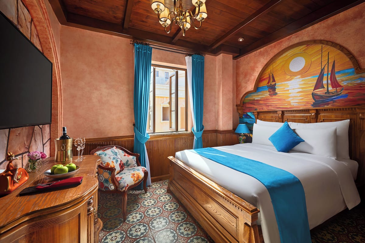 Une chambre d’hôtel avec un lit en bois, un bureau sur lequel se trouvent une bouteille et des pommes, une chaise, et une vue sur l’extérieur par une fenêtre ornée de rideaux bleus.
