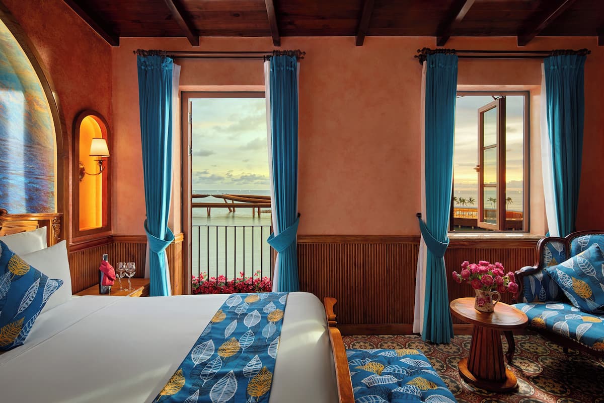 Une chambre d’hôtel avec un lit, un canapé, une table ornée d’un vase de fleurs et une vue sur la mer grâce à de grandes fenêtres.