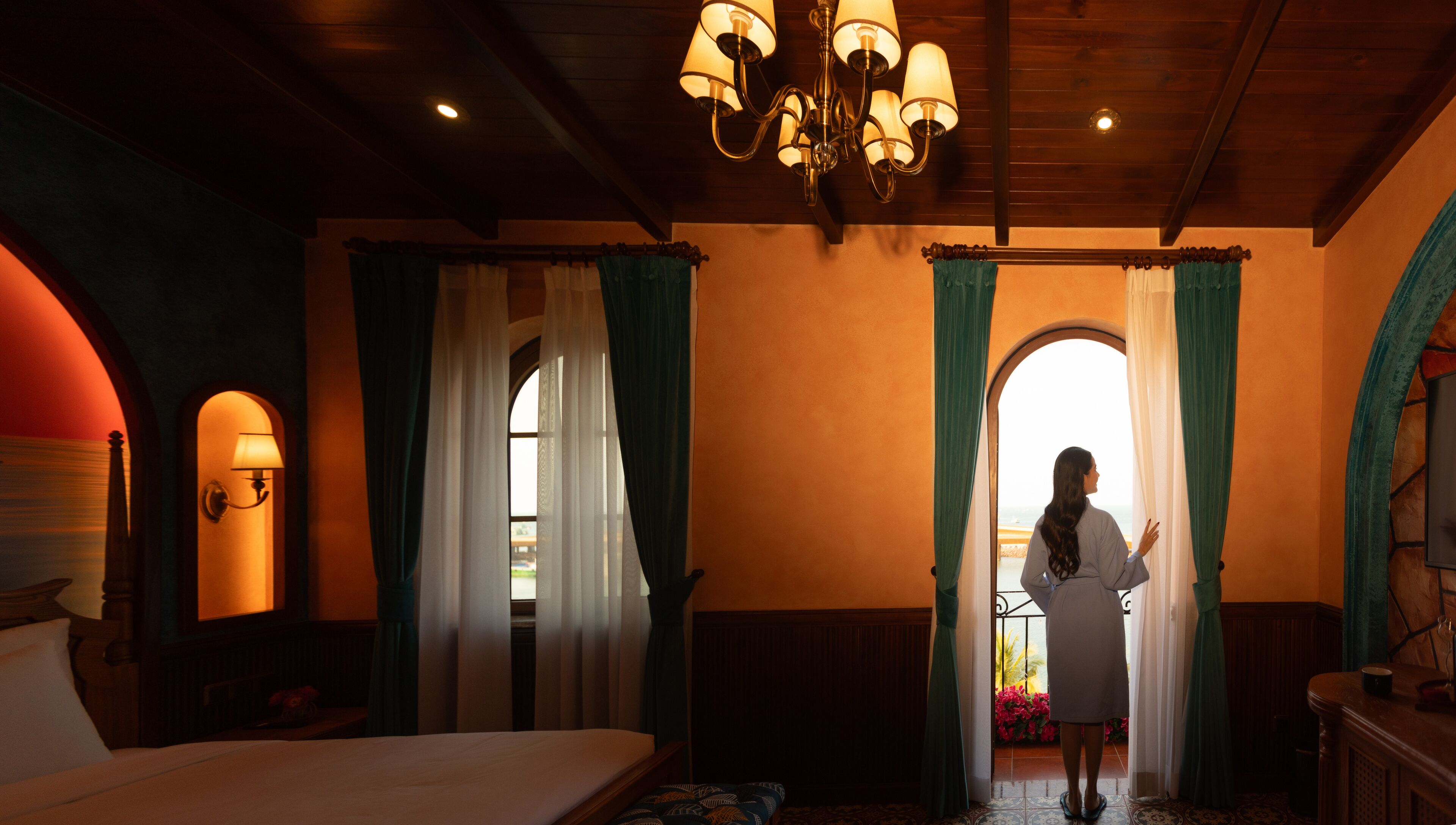 A hotel room with a bed, a chandelier, curtains, and a view of the outdoors.