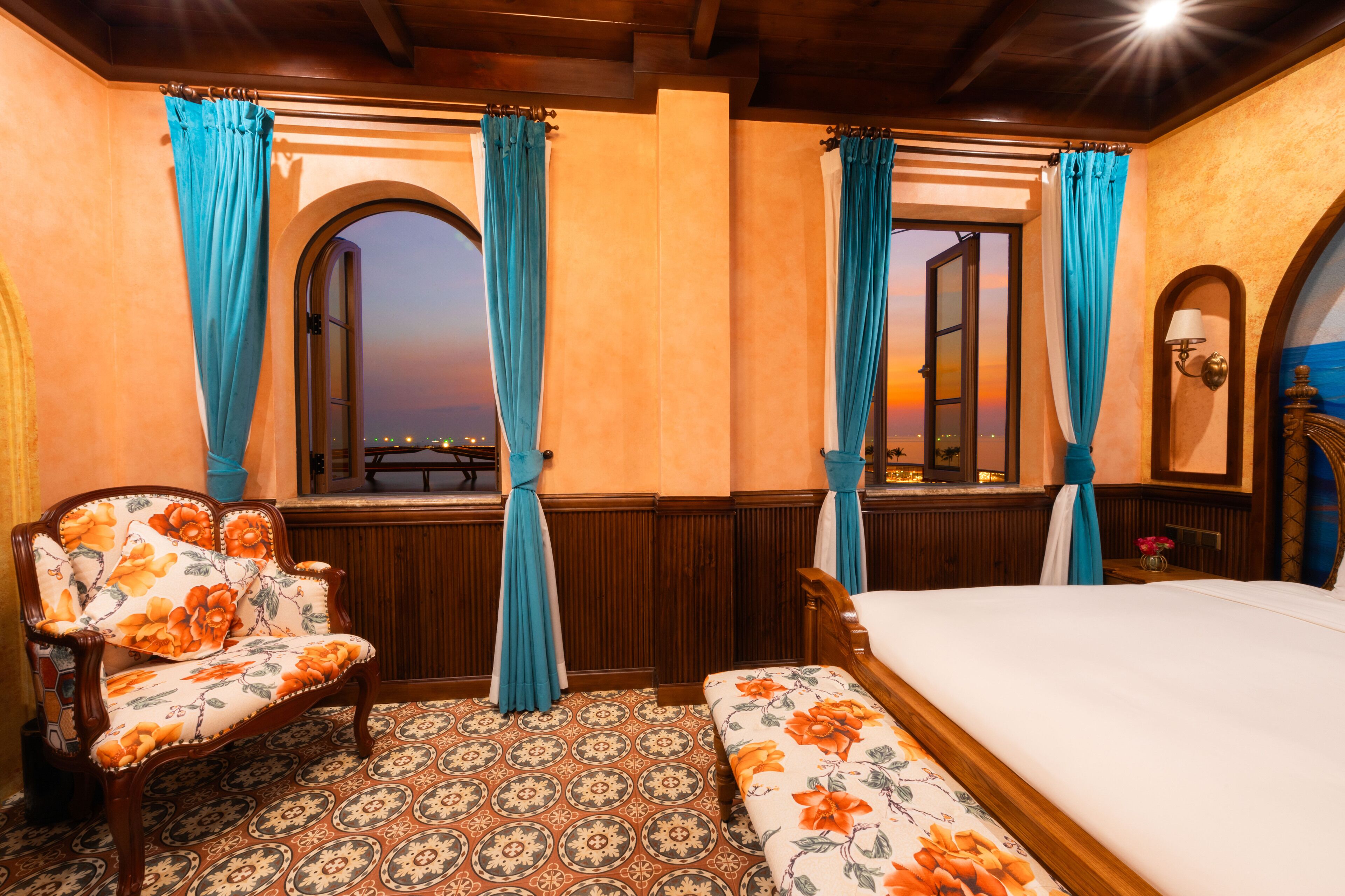 A room with a bed, a bench, and a chair with floral upholstery. There are two windows with curtains, and a view of the cityscape at sunset.