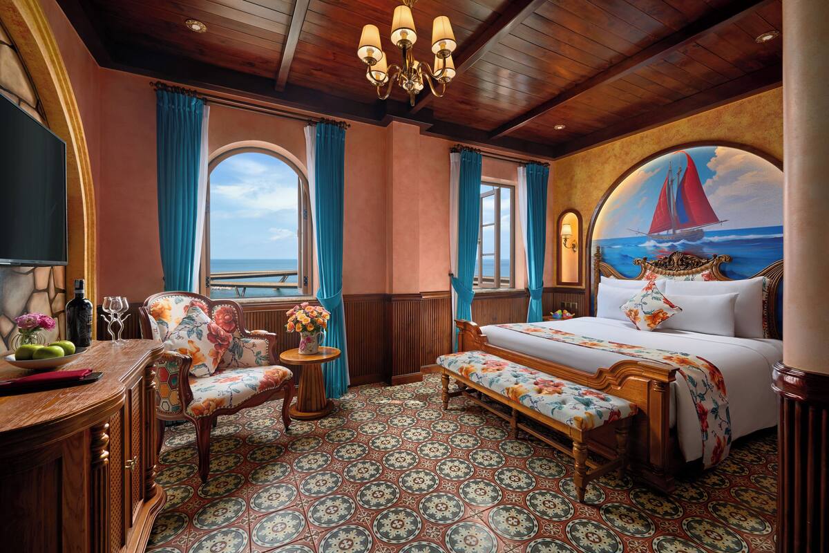 Une chambre avec un lit, un bureau, une chaise, un banc et une vue sur la mer par la fenêtre.