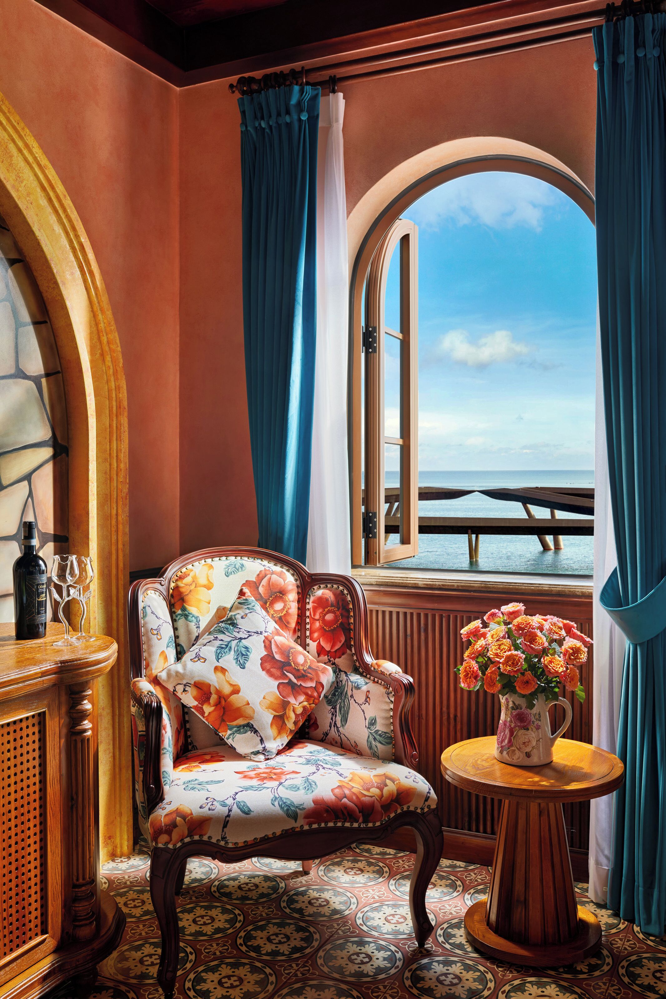Una stanza con vista sul mare, una sedia in legno con rivestimento floreale, un tavolo con un vaso di fiori e una bottiglia e dei bicchieri su un tavolino.
