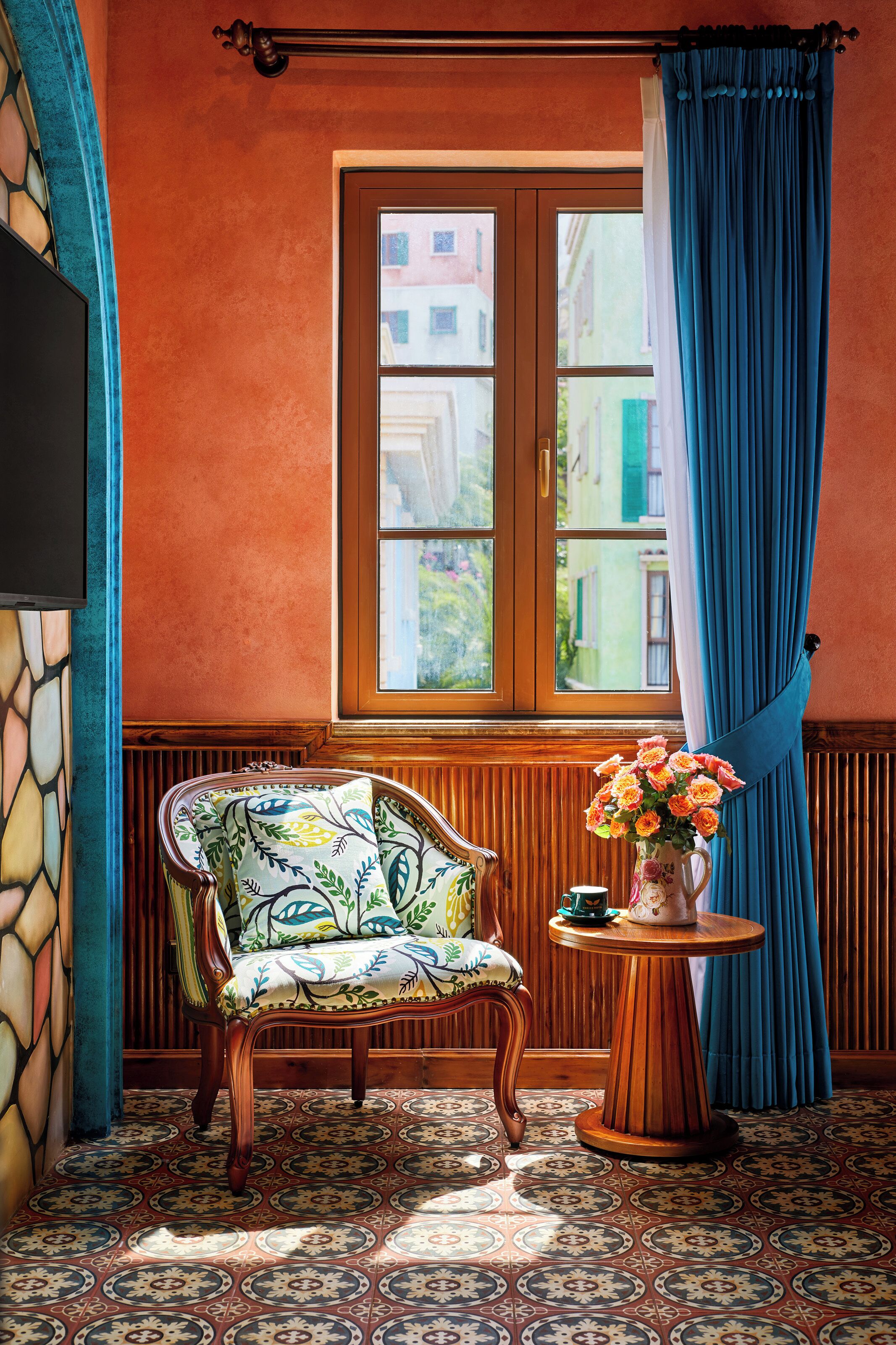 Une pièce avec un sol à motifs, une chaise en bois, une table ornée d’un vase de fleurs et une fenêtre avec des rideaux bleus.