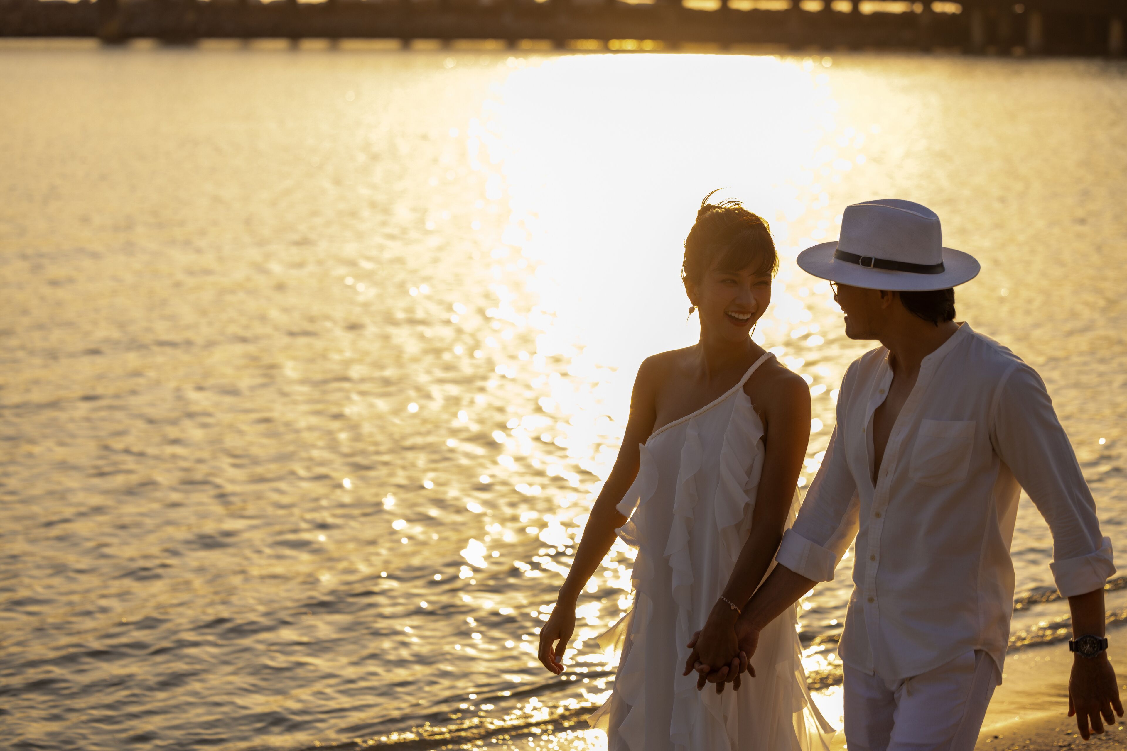A couple walking hand in hand by a body of water during sunset.