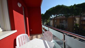 En balkon med et hvidt bord og stole, en rød væg samt udsigt til omkringliggende bygninger og træer.