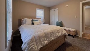 A bedroom with a large bed, a nightstand, a dresser, and a window with blinds. - Perfect for families! (Newport News)