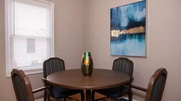 A dining room with a round table, six chairs, a vase, and a painting on the wall.