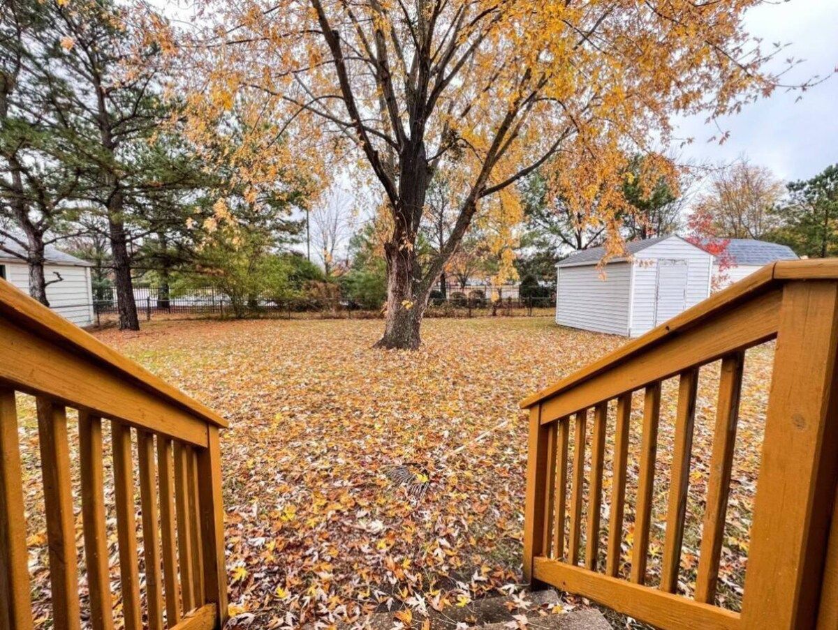 A wooden deck with a view of a yard covered in fallen leaves, a tree with yellow leaves, and a white building in the background.