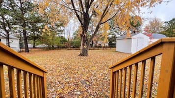 A wooden deck with a view of a yard covered in fallen leaves, a tree with yellow leaves, and a white building in the background.