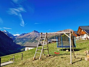 Un parc infantil amb balancí i escala, envoltat de cases i muntanyes.