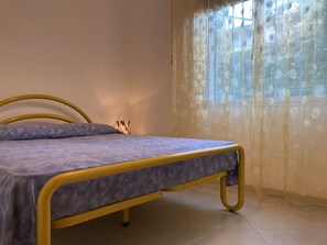 Un lit simple avec un cadre jaune, un matelas gris et une fenêtre ornée de rideaux à motifs floraux.