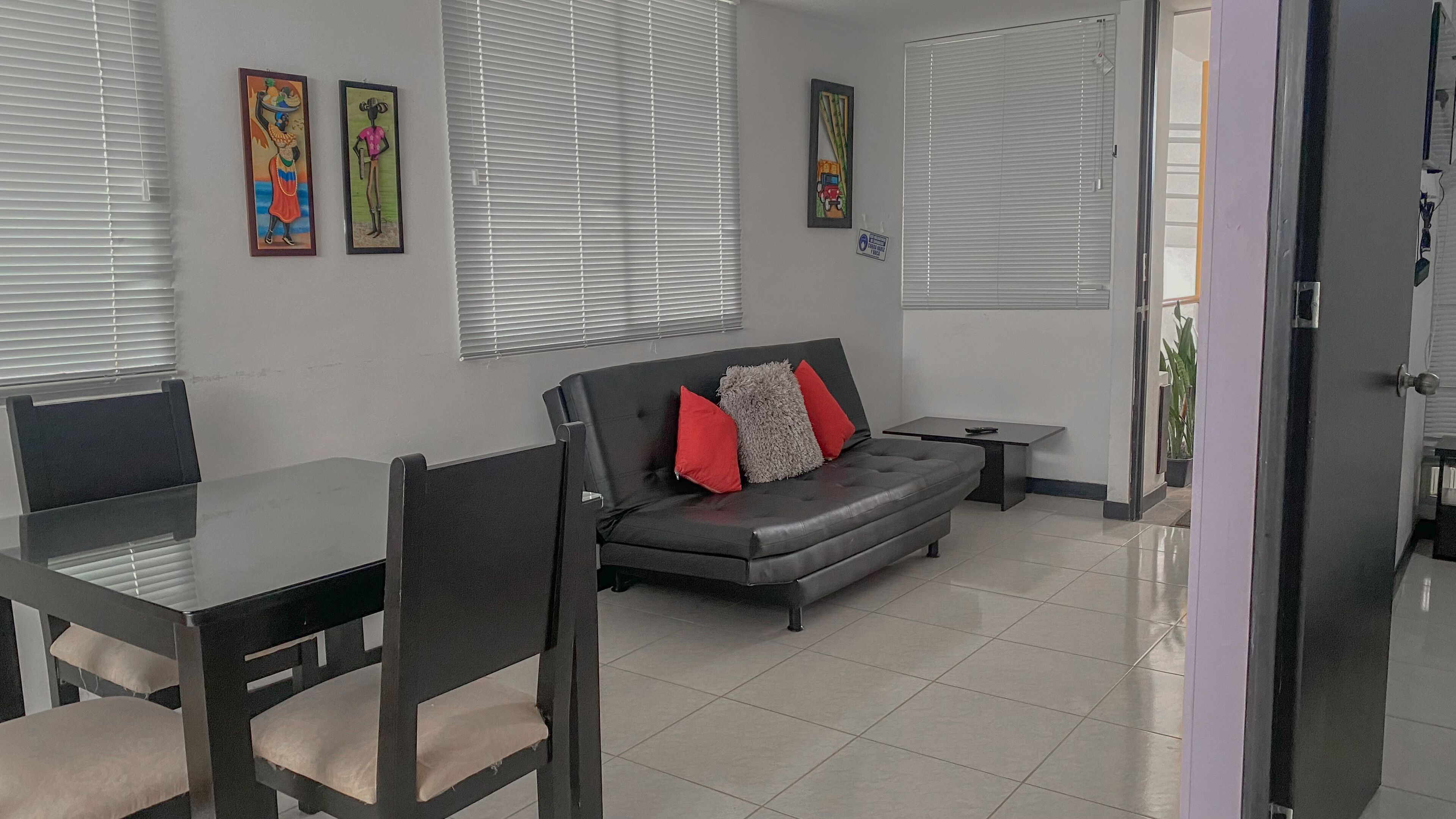 Una sala de estar moderna con un sofá, una mesa de comedor y sillas. Hay dos ventanas con persianas, una mesita auxiliar y obras de arte en las paredes.
