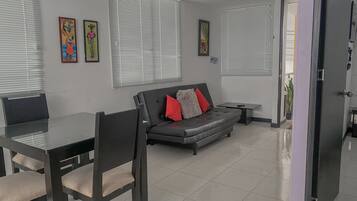 Una sala de estar moderna con un sofá, una mesa de comedor y sillas. Hay dos ventanas con persianas, una mesita auxiliar y obras de arte en las paredes.