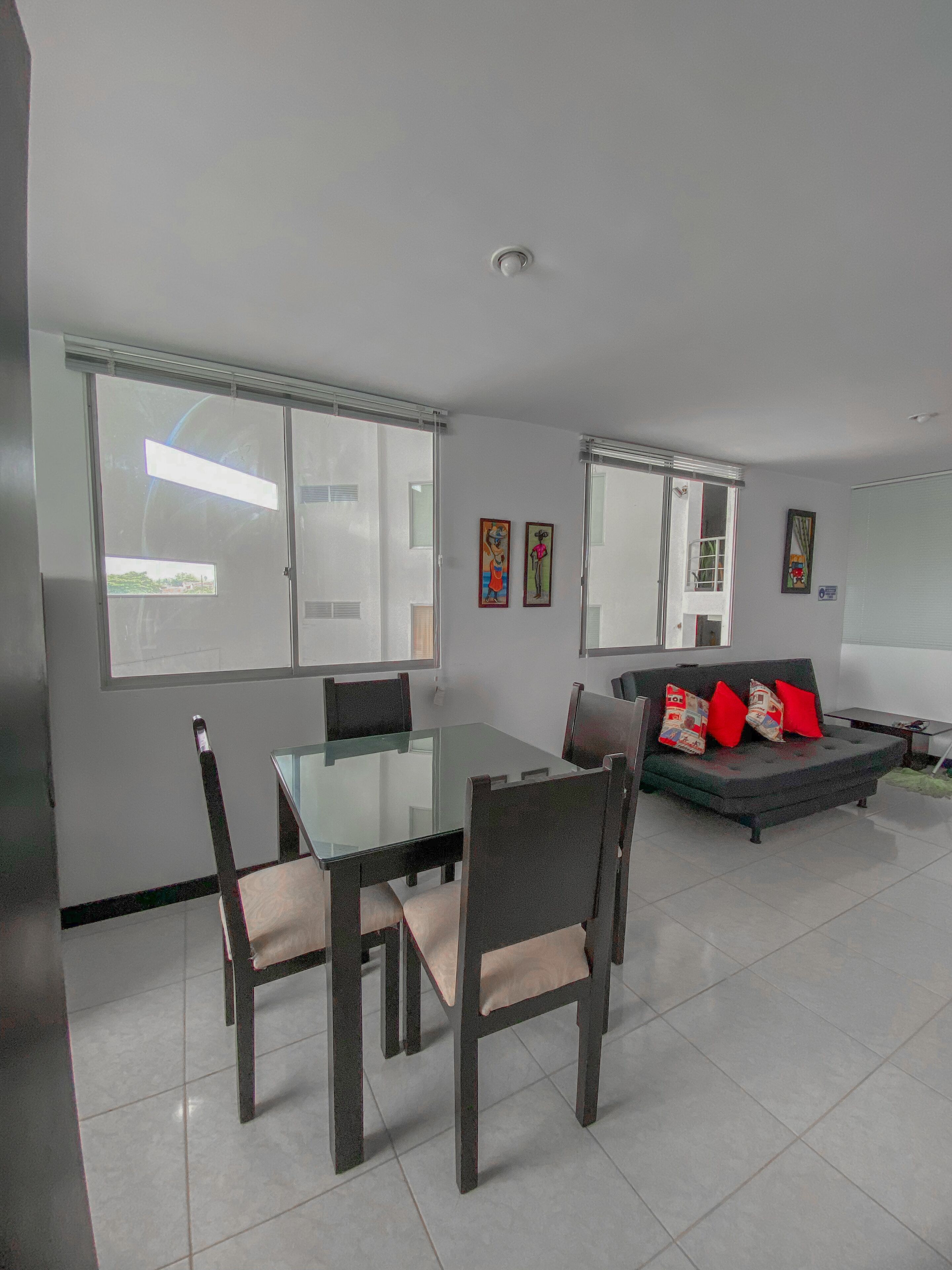 A modern living room with a glass dining table, dark wooden chairs, a grey sofa with red cushions, and a large window providing natural light.