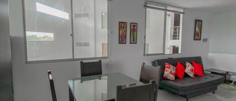A modern living room with a glass dining table, dark wooden chairs, a grey sofa with red cushions, and a large window providing natural light.