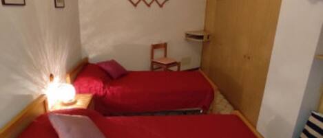 A small, cozy room with two beds, a wooden wardrobe, a chair, and a small table.