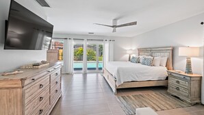 Quarto com cama, televisão na parede, ventilador de teto e vista para uma piscina através das portas de vidro.