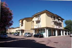 A multi-story building with balconies, a paved ground floor, and parked cars.