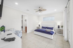 A bedroom with a bed, bedside tables, a ceiling fan, a window with decorative glass, and a clock on the wall. - Coastal Two-Story Home | On Water w/ Pool/Dock (Marco Island)