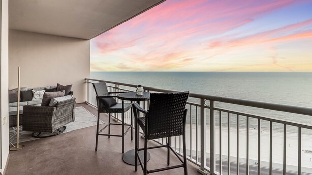Un balcon avec vue sur la mer et sur le coucher de soleil. Il y a deux chaises et une table.