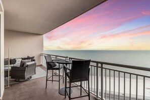 Un balcon avec vue sur la mer et sur le coucher de soleil. Il y a deux chaises et une table.