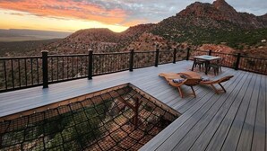 A wooden deck with a table and chairs, overlooking a mountainous landscape during sunset.