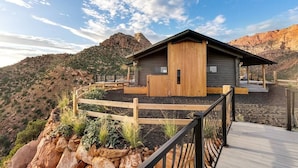 A cabin with a wooden deck, surrounded by a wooden fence, set against a mountainous backdrop.