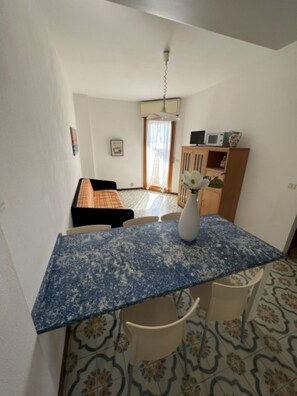 A small, compact room with a kitchenette, a dining area with a blue patterned table, and a bed in the background. - Seaside bliss - Beahost Rentals (Bibione)